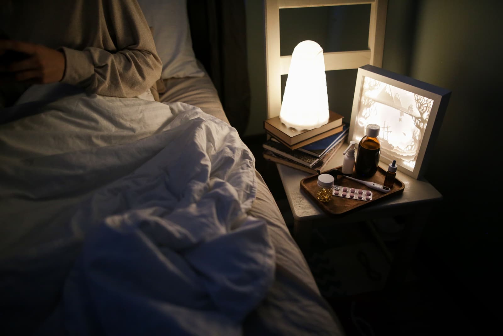 A bedside lamp lit low beside books, a bottle and a tray of tablets.